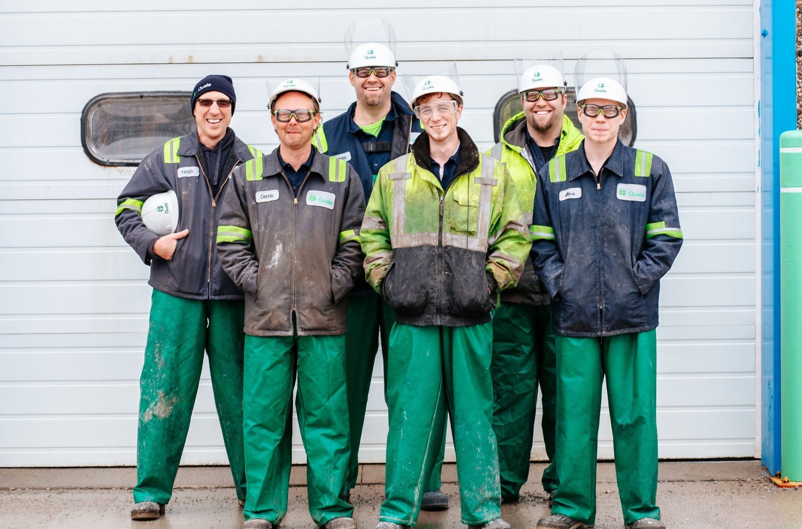 Six industrial workers in safety gear smiling in front of a garage door.